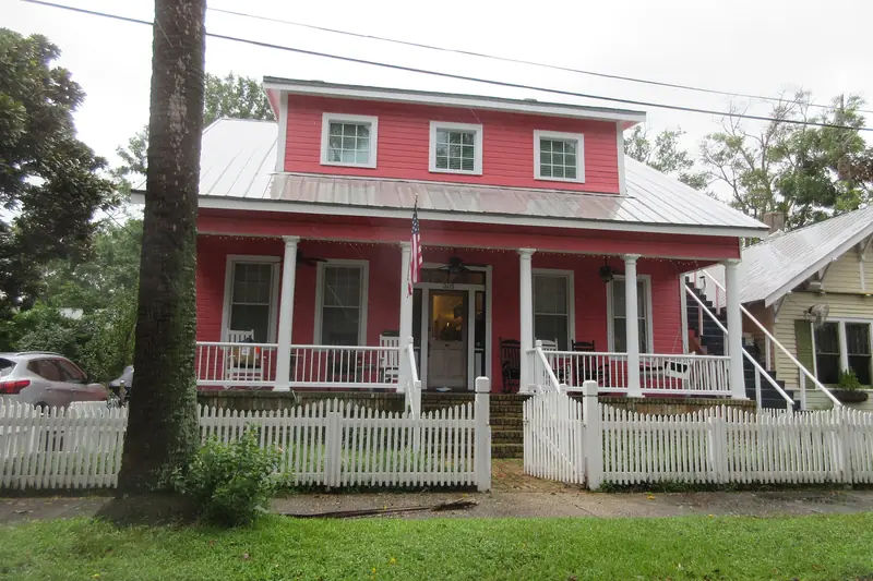 Coral-colored raised house with a deep shaded porch, fans spinning on the porch ceiling and a pale metal roof behind a white picket fence